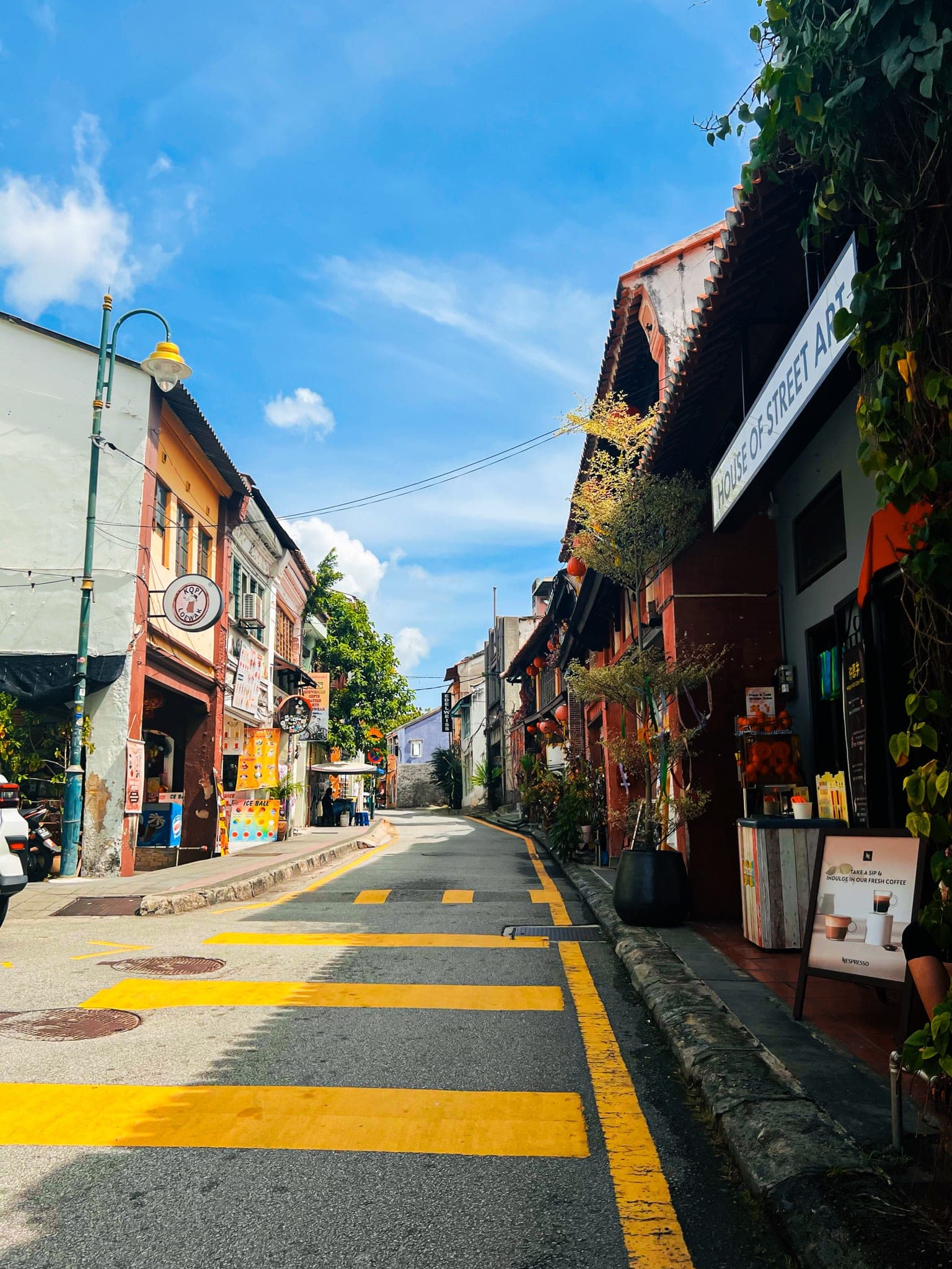 A sunlit George Town street lined with colorful shophouses, with bright yellow road markings under a blue sky