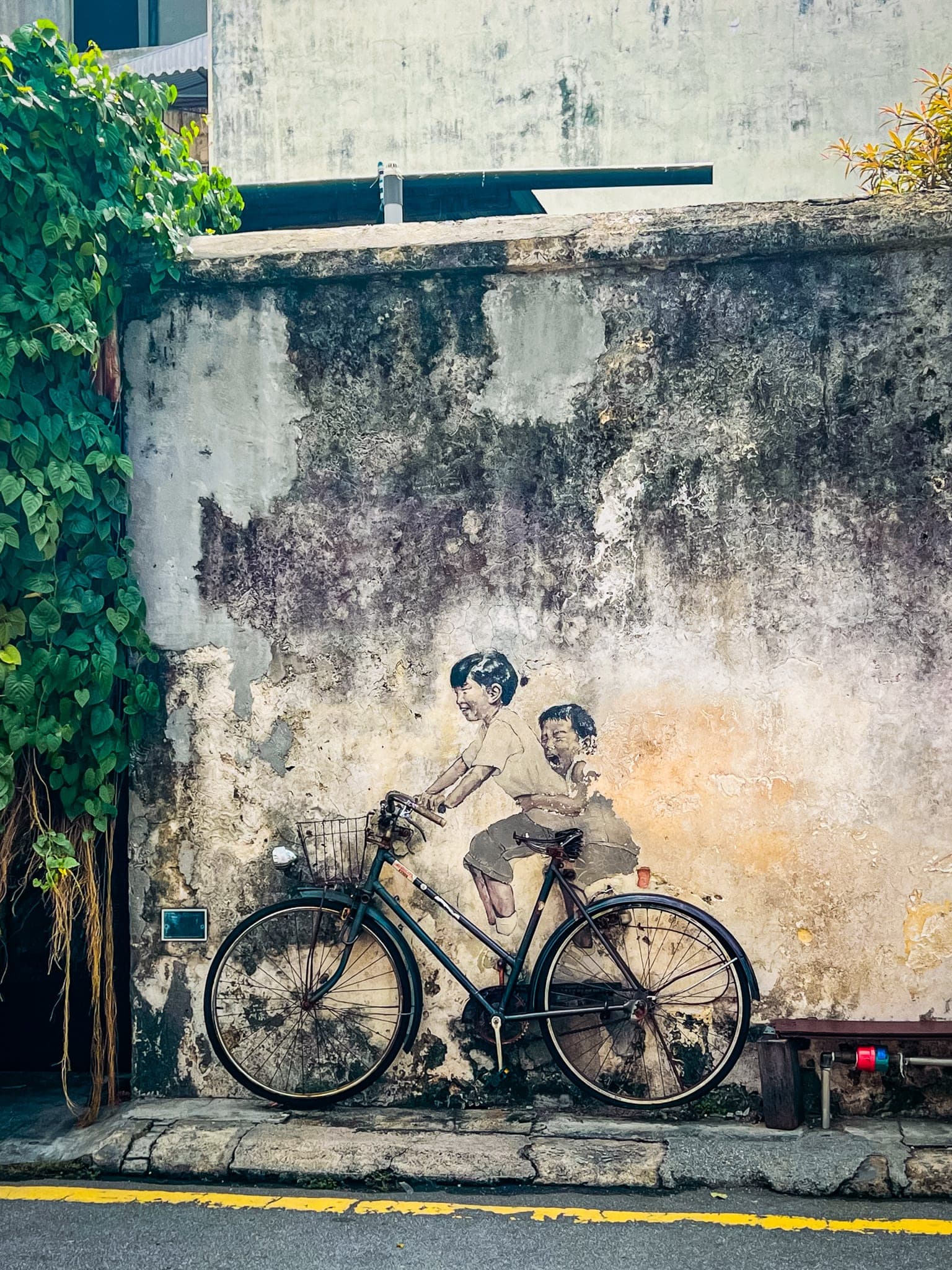 A mural of two children painted on a weathered wall, riding a real bicycle fixed in front of it